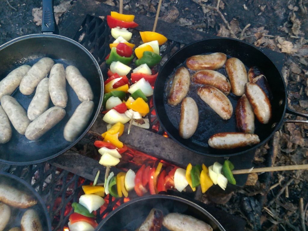 Sausages sizzling in a pan and skewered veg cooking over the open fire
